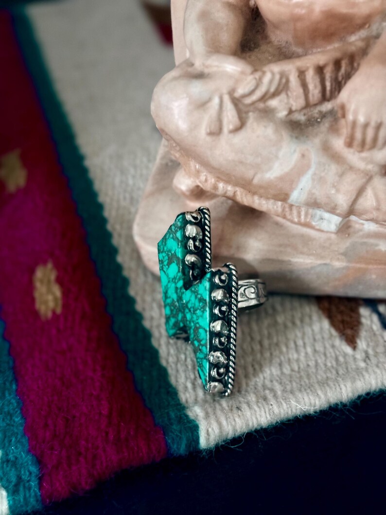 May include: A silver ring with a turquoise stone in a lightning bolt shape. The turquoise is a vibrant green-blue color. The ring has a detailed, textured design around the stone and on the band.