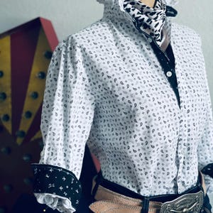 Peut inclure: Chemise blanche &agrave; motifs cachemire, bordures noires et poignets avec un motif d'avion. La chemise a un col &agrave; volants et est associ&eacute;e &agrave; une &eacute;charpe &agrave; motifs, une ceinture marron clair et une boucle argent&eacute;e.