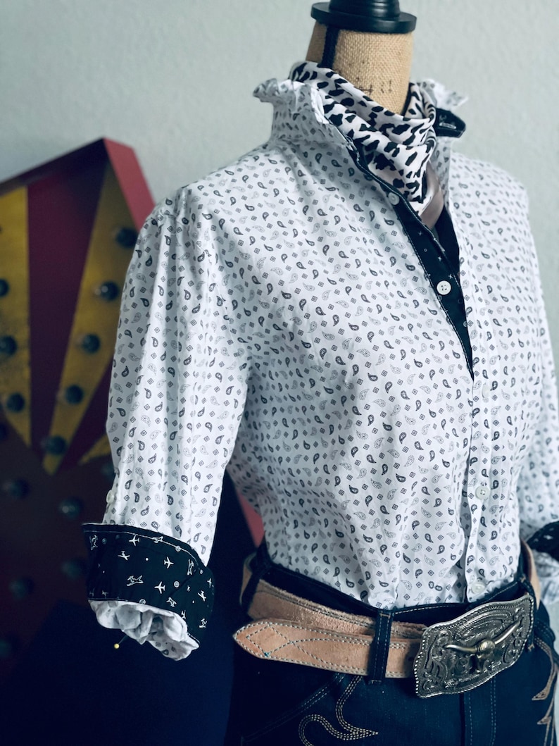 Peut inclure: Chemise blanche &agrave; motifs cachemire, bordures noires et poignets avec un motif d'avion. La chemise a un col &agrave; volants et est associ&eacute;e &agrave; une &eacute;charpe &agrave; motifs, une ceinture marron clair et une boucle argent&eacute;e.