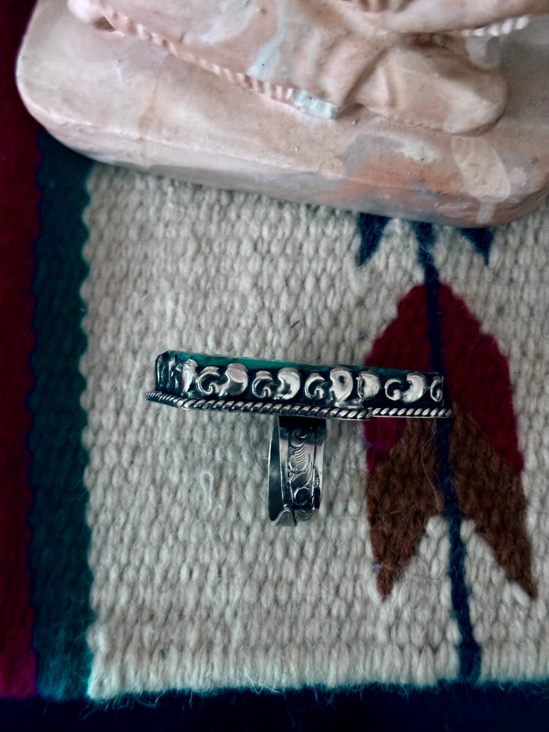May include: A silver ring with a rectangular, turquoise-colored stone. The ring has a detailed, ornate design around the stone and on the band. The ring is displayed on a patterned textile with a Native American design.