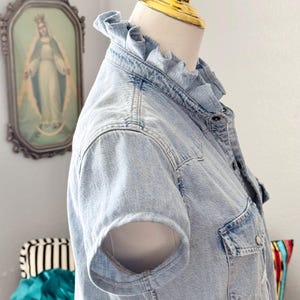 Peut inclure: Chemise en jean bleu clair &agrave; manches courtes et col &agrave; volants. La chemise a une fermeture boutonn&eacute;e sur le devant, une poche poitrine et un design en jean classique. La chemise est pr&eacute;sent&eacute;e sur un mannequin.