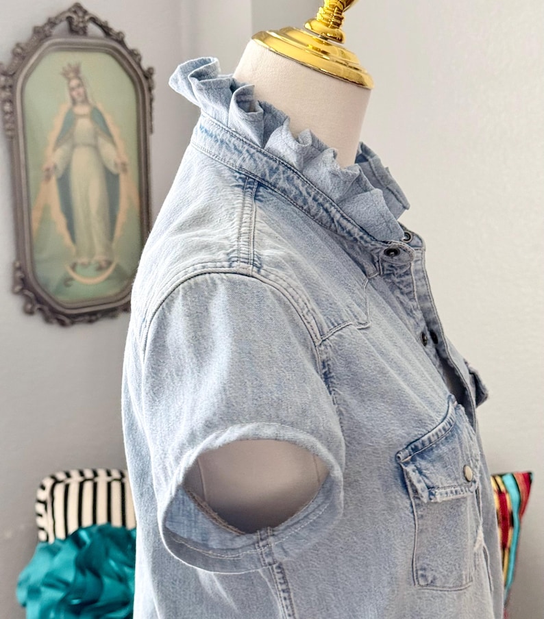 Peut inclure: Chemise en jean bleu clair &agrave; manches courtes et col &agrave; volants. La chemise a une fermeture boutonn&eacute;e sur le devant, une poche poitrine et un design en jean classique. La chemise est pr&eacute;sent&eacute;e sur un mannequin.