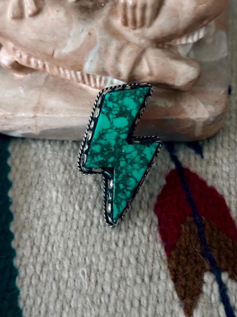 May include: A turquoise-colored lightning bolt-shaped pendant with a silver-colored rope-like border. The turquoise has dark green veining. The pendant is set against a textured, off-white background.