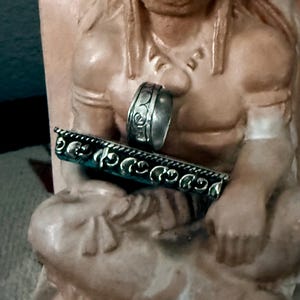 May include: A silver ring with an engraved floral design and a silver bar with a turquoise background and a decorative edge. The jewelry is displayed on a statue of a Native American.