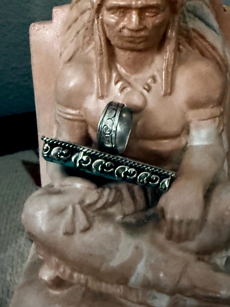 May include: A silver ring with an engraved floral design and a silver bar with a turquoise background and a decorative edge. The jewelry is displayed on a statue of a Native American.