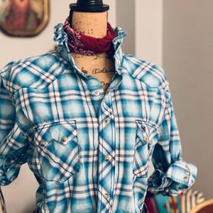 Peut inclure: Une chemise western &agrave; carreaux bleus et blancs avec des fermetures &agrave; boutons-pression et des poches. La chemise est pr&eacute;sent&eacute;e sur un mannequin avec un bandana rouge autour du cou. Une ceinture rose avec une grosse boucle est &eacute;galement visible.