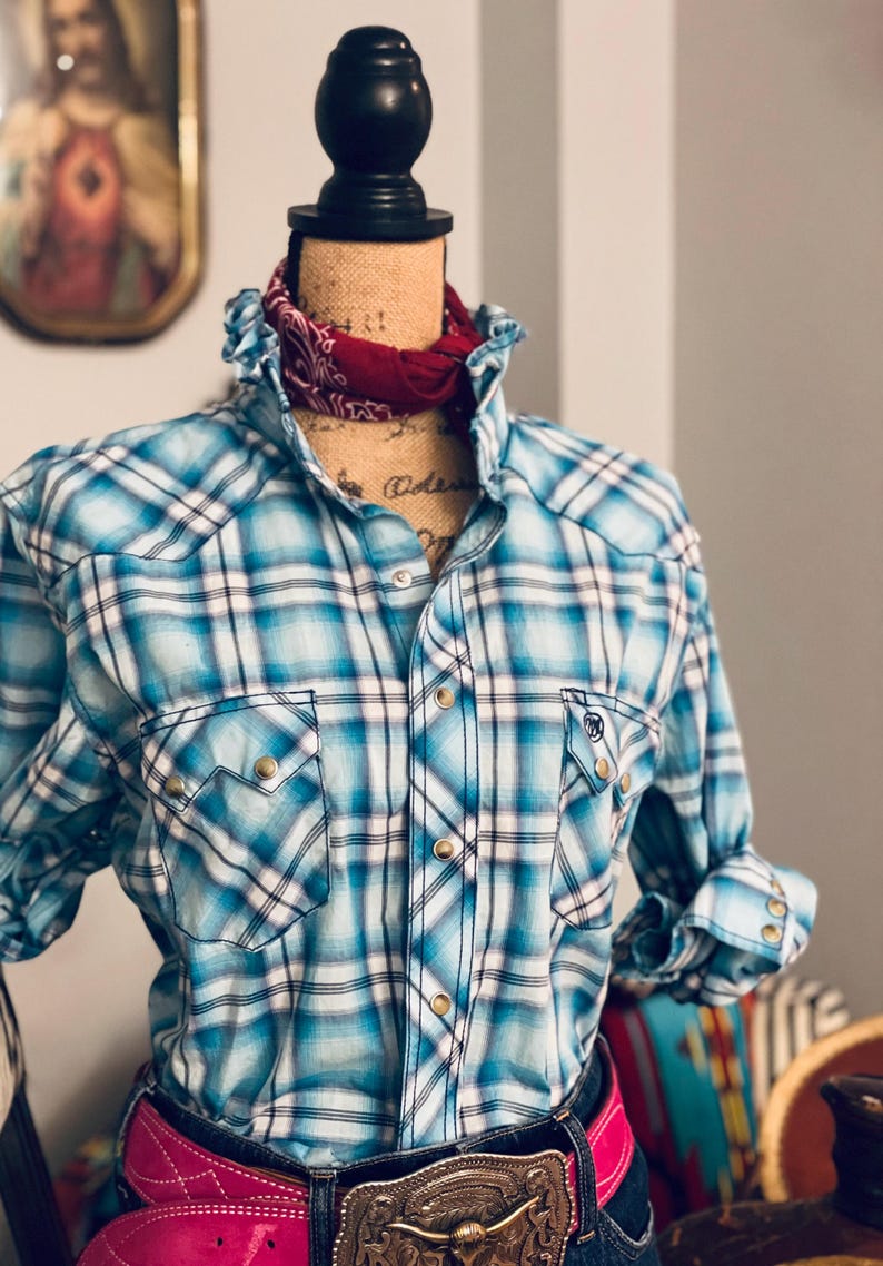 Peut inclure: Une chemise western &agrave; carreaux bleus et blancs avec des fermetures &agrave; boutons-pression et des poches. La chemise est pr&eacute;sent&eacute;e sur un mannequin avec un bandana rouge autour du cou. Une ceinture rose avec une grosse boucle est &eacute;galement visible.