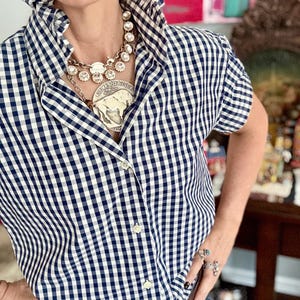 Peut inclure: Une chemise &agrave; carreaux vichy bleu marine et blanc &agrave; manches courtes et grand col. La chemise est boutonn&eacute;e et port&eacute;e avec un grand collier argent&eacute;. La personne porte &eacute;galement des bagues et une ceinture &agrave; boucle d&eacute;corative. Style classique et d&eacute;contract&eacute;.