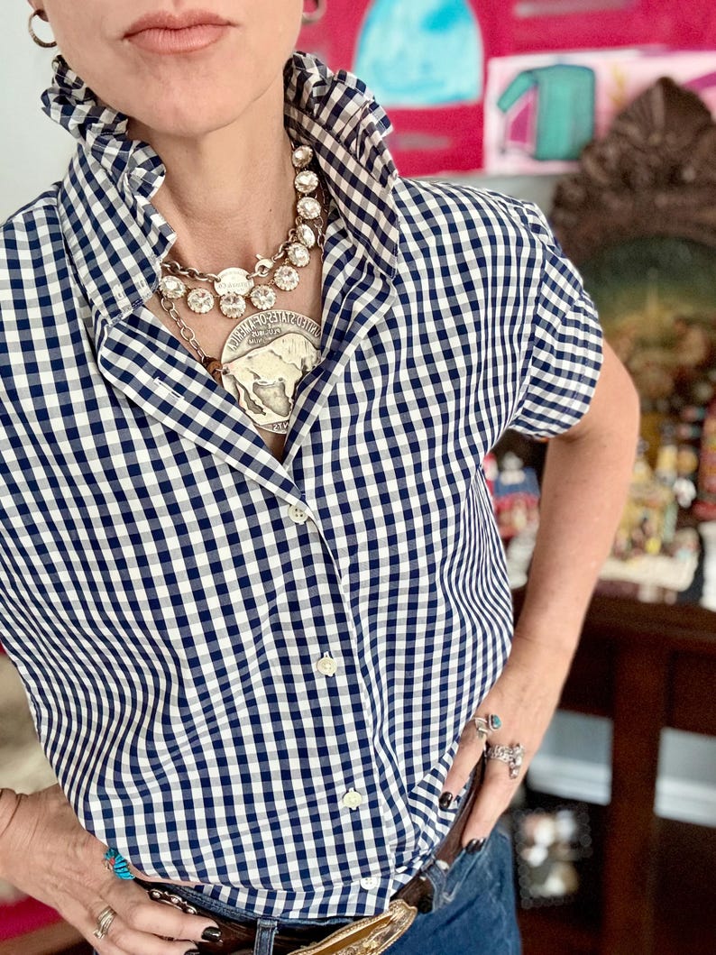 Peut inclure: Une chemise &agrave; carreaux vichy bleu marine et blanc &agrave; manches courtes et grand col. La chemise est boutonn&eacute;e et port&eacute;e avec un grand collier argent&eacute;. La personne porte &eacute;galement des bagues et une ceinture &agrave; boucle d&eacute;corative. Style classique et d&eacute;contract&eacute;.