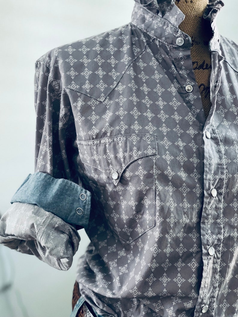 Peut inclure: Chemise grise &agrave; boutons avec un motif g&eacute;om&eacute;trique blanc. La chemise a deux poches avant, un col et les manches sont retrouss&eacute;es pour r&eacute;v&eacute;ler un poignet en jean. La chemise est pr&eacute;sent&eacute;e sur un mannequin.