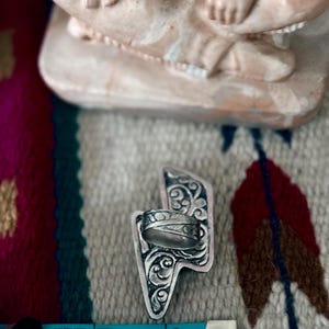 May include: A silver ring with an engraved design, set on a decorative silver lightning bolt-shaped base. The ring is positioned near a blue measuring tape, showing inches. The background features a patterned textile and a beige figurine.