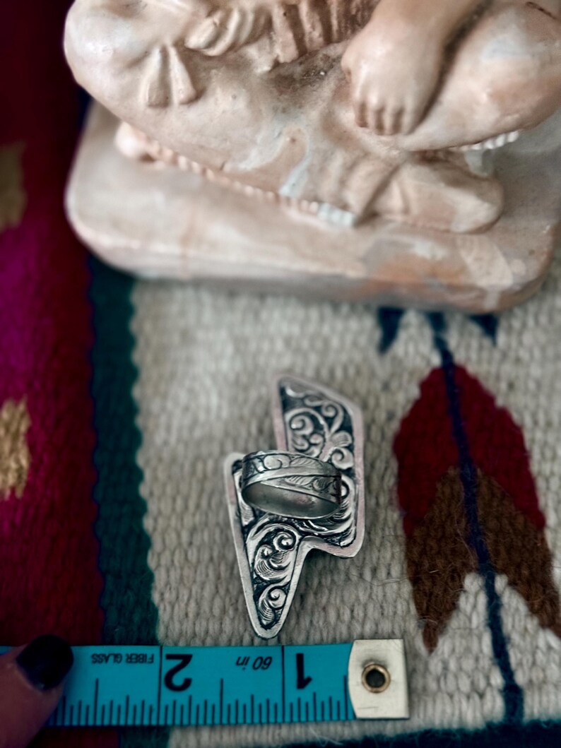 May include: A silver ring with an engraved design, set on a decorative silver lightning bolt-shaped base. The ring is positioned near a blue measuring tape, showing inches. The background features a patterned textile and a beige figurine.