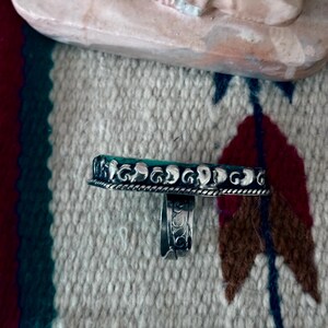 May include: A silver ring with a rectangular, turquoise-colored stone. The ring has a detailed, ornate design around the stone and on the band. The ring is displayed on a patterned textile with a Native American design.