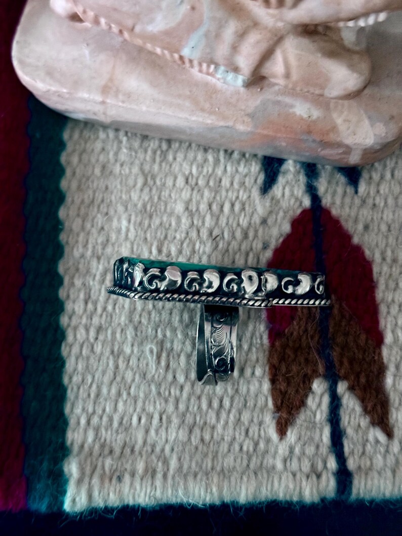May include: A silver ring with a rectangular, turquoise-colored stone. The ring has a detailed, ornate design around the stone and on the band. The ring is displayed on a patterned textile with a Native American design.
