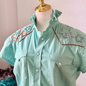 May include: A light blue short-sleeved shirt with a vertical striped pattern and small black dots. The shirt features star-shaped embroidery with brown trim on the shoulders and chest pockets. It has a ruffled collar and pearlized buttons.