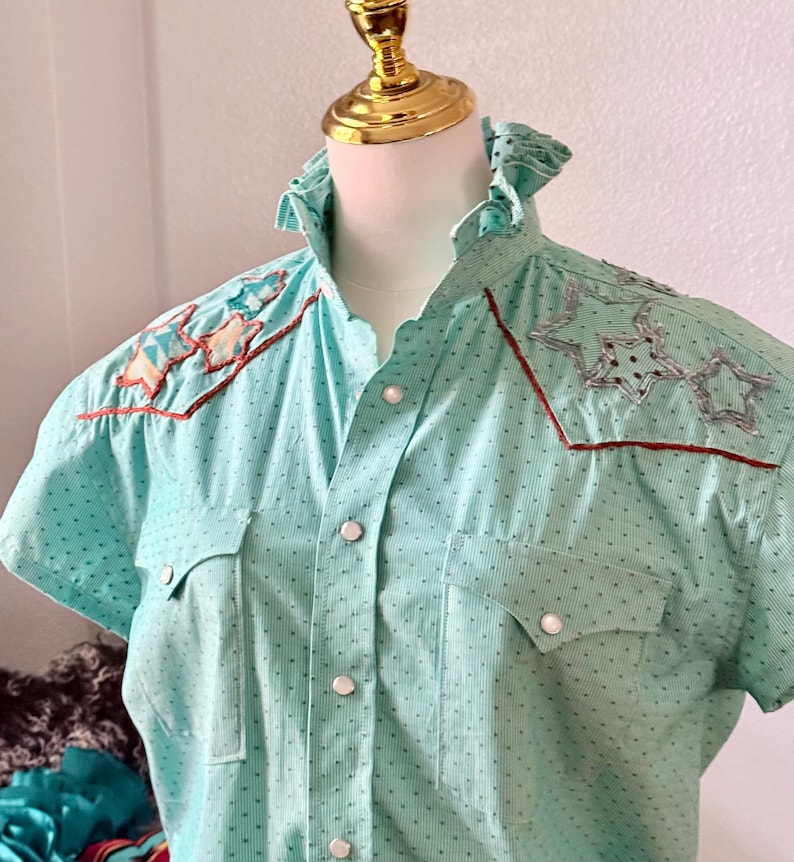 May include: A light blue short-sleeved shirt with a vertical striped pattern and small black dots. The shirt features star-shaped embroidery with brown trim on the shoulders and chest pockets. It has a ruffled collar and pearlized buttons.