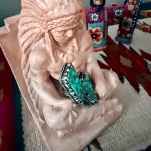 May include: A pair of turquoise-colored gemstone rings with silver-colored metal settings. The rings are resting on a light pink statue of a Native American chief. The statue is in a seated position. Other decorative items are in the background.