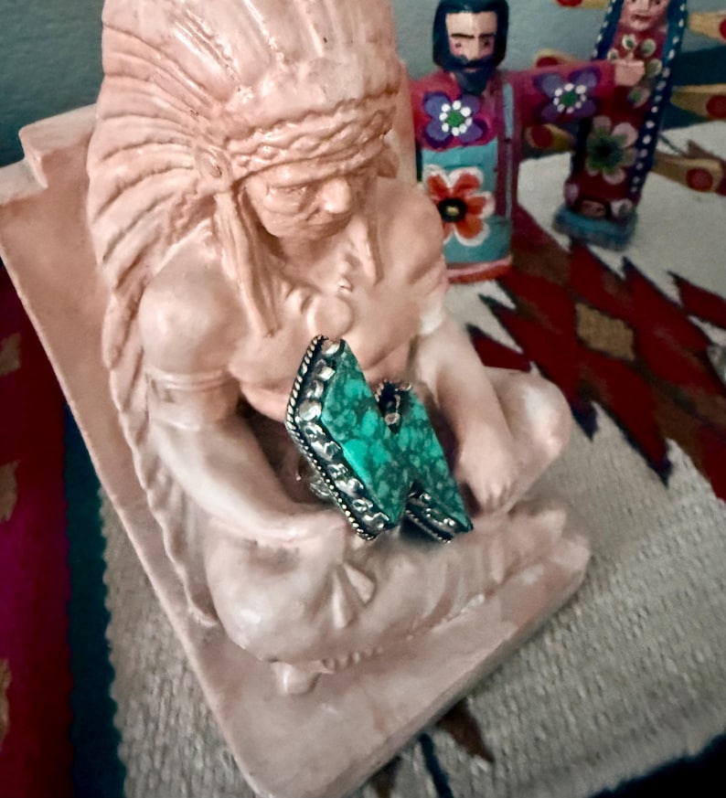 May include: A pair of turquoise-colored gemstone rings with silver-colored metal settings. The rings are resting on a light pink statue of a Native American chief. The statue is in a seated position. Other decorative items are in the background.