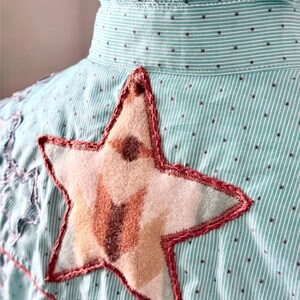 May include: Close-up of a light blue and white striped shirt with a large star applique. The star is made of beige and brown fabric with a red stitched outline. The shirt has a ruffled collar and small black polka dots.