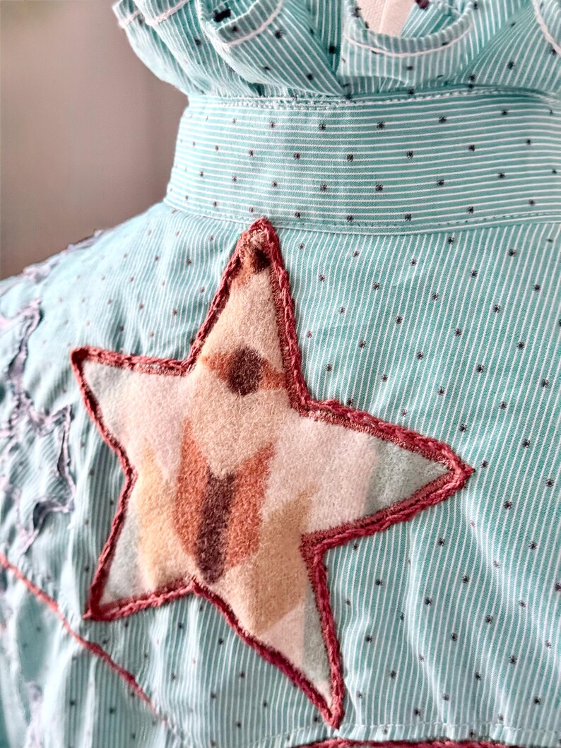 May include: Close-up of a light blue and white striped shirt with a large star applique. The star is made of beige and brown fabric with a red stitched outline. The shirt has a ruffled collar and small black polka dots.