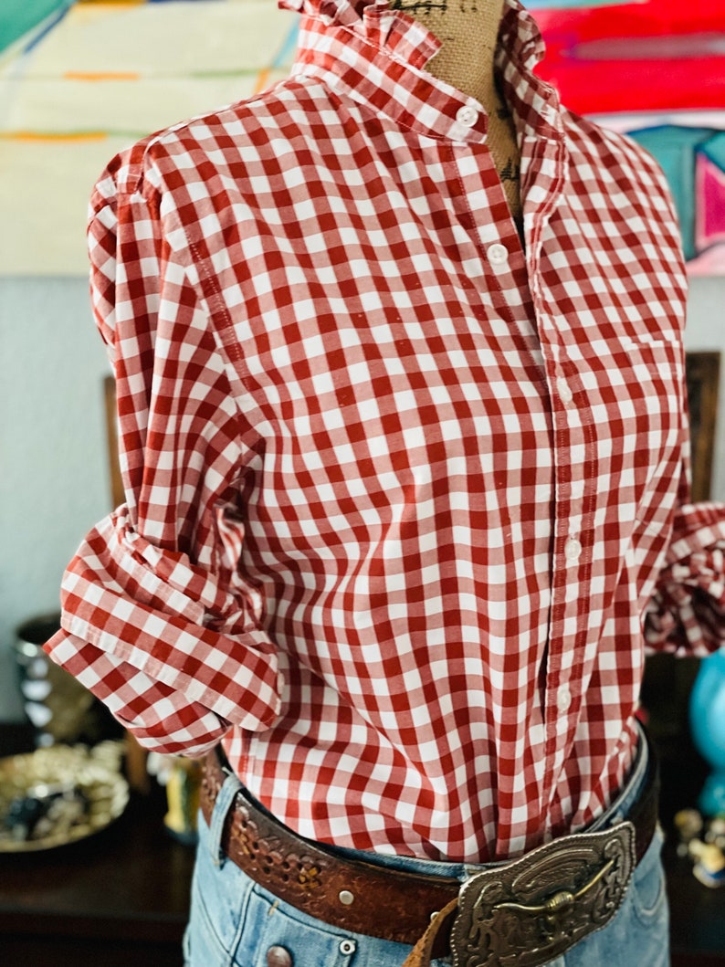 Peut inclure: Chemise &agrave; carreaux rouges et blancs avec un col &agrave; volants et une fermeture boutonn&eacute;e. La chemise &agrave; manches longues est associ&eacute;e &agrave; un jean bleu et une ceinture en cuir marron avec une boucle argent&eacute;e d&eacute;corative. Les manches sont retrouss&eacute;es.