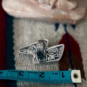 May include: A silver ring with an ornate, stylized design. The ring's decorative elements include swirling patterns. A blue measuring tape shows the ring is approximately 1.5 inches in length.