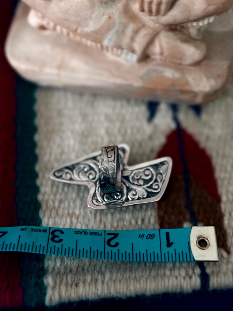 May include: A silver ring with an ornate, stylized design. The ring's decorative elements include swirling patterns. A blue measuring tape shows the ring is approximately 1.5 inches in length.