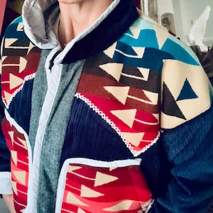 May include: A jacket featuring a mix of navy blue corduroy, grey herringbone fabric, and panels with a colorful geometric arrow pattern in red, brown, cream, and teal. The garment includes a high collar, white trim, and two front pockets.
