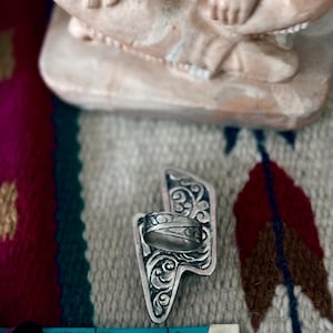 May include: A silver ring with an engraved design, set on a decorative silver lightning bolt-shaped base. The ring is positioned near a blue measuring tape. The background features a patterned textile and a beige figurine.