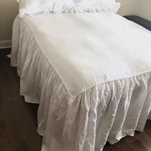May include: White linen bed skirt with a ruffled edge. The skirt is on a bed with two white pillowcases with ruffled edges.