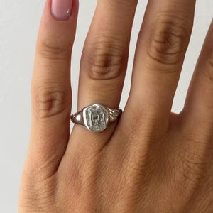 Könnte beinhalten: Ein silberfarbener Ring mit einem rechteckigen, facettierten, klaren Stein, der in eine dicke, organisch geformte Metallschiene eingefasst ist. Die Fassung hat eine glatte, polierte Oberfläche mit einer leicht unregelmäßigen, abgerundeten Kontur.