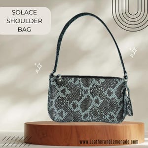 Könnte beinhalten: Eine Solace-Umhängetasche mit Schlangenmuster in Grau- und Türkistönen. Die Tasche hat einen schwarzen Reißverschluss, einen passenden Griff und eine kleine Quaste. Die Tasche wird auf einem Holzständer präsentiert.