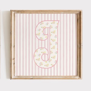 May include: A pink and white floral letter J on a pink and white striped background. The letter is in a decorative font and is framed in a light wood frame.