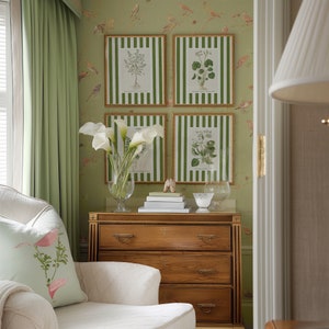 May include: A white upholstered armchair with a green floral pillow sits in front of a wooden dresser with three drawers. The dresser is against a green wall with four framed botanical prints.
