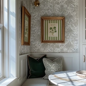 May include: A white and gray patterned wallpaper with two gold framed botanical prints. A white bench with green and gray patterned pillows sits in the corner of the room. A white marble table with a gold rim sits in front of the bench.