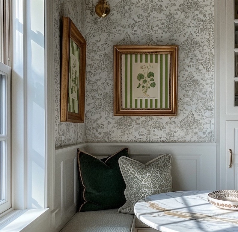 May include: A white and gray patterned wallpaper with two gold framed botanical prints. A white bench with green and gray patterned pillows sits in the corner of the room. A white marble table with a gold rim sits in front of the bench.