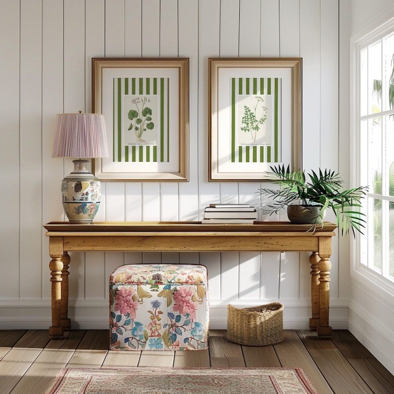 May include: A wooden console table with a floral patterned ottoman, a wicker basket, and a lamp with a pink shade. Two framed botanical prints with green and white stripes hang on the wall behind the table.