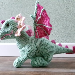 May include: A crocheted green dragon toy with pink wings and a pink and gold flower on its head.