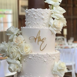 May include: A three-tiered white wedding cake decorated with pearl-like accents and white roses. The top tier has the names "A & E" written in gold script.
