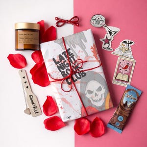 May include: A still life arrangement with a wrapped book, candle, and decorative items. The book is wrapped in white paper with a skull graphic and tied with red twine. The text "LATE NIGHT BOOK CLUB" is visible. Rose petals and stickers complete the scene.