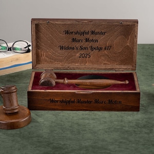 Könnte beinhalten: Ein hölzerner Hammer und ein Schallblock liegen neben einer offenen Holzkiste mit einem Hammer und einem Schallblock. Die Kiste ist mit Text graviert. Ein Buch und eine Brille sind im Hintergrund. Der Hammer ist ein Symbol der Autorität.