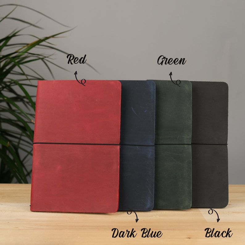 Peut inclure: Quatre journaux reli&eacute;s en cuir de couleur rouge, bleu fonc&eacute;, vert et noir. Les journaux sont munis de fermetures &eacute;lastiques et sont dispos&eacute;s en rang&eacute;e sur une surface en bois.