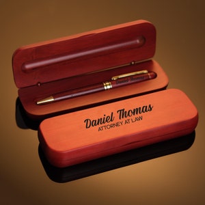 May include: A mahogany wood pen box with a gold pen inside. The box is engraved with the text "Daniel Thomas Attorney at Law".