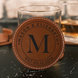 Könnte beinhalten: Ein brauner Lederuntersetzer mit weißen Nähten zeigt die Namen "Mason & Elizabeth" und den Buchstaben "M" in einem Kreis. Der Text "EST. 20.12.2018" ist ebenfalls enthalten. Zwei Gläser sind im Hintergrund.