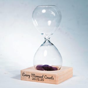 May include: A clear glass hourglass with purple sand, resting on a light-colored wooden base. The base is engraved with the words "Every Moment Counts" and the date "20.12.25". The hourglass is a decorative item.