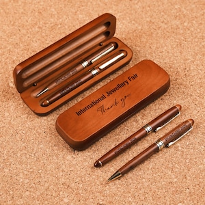 May include: A set of two wooden pens and a pencil, presented in a wooden case. The pens and pencil are dark brown with silver accents. The case is engraved with "VICENZAORO 2025" and "International Jewellery Fair Thank you."