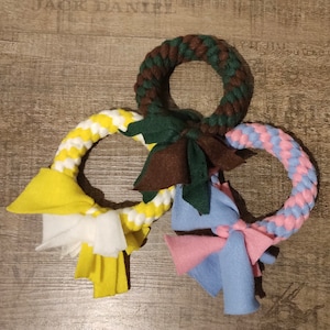Könnte beinhalten: Drei Hundespielzeuge in Form von Ringen, aus geflochtenem Seil und Stoff. Die Ringe sind in Gelb und Weiß, Grün und Braun sowie Rosa und Blau.