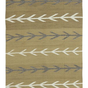 Puede incluir: Una alfombra tejida de color marrón claro y blanco con un patrón geométrico de flechas. La alfombra tiene un borde negro y blanco.