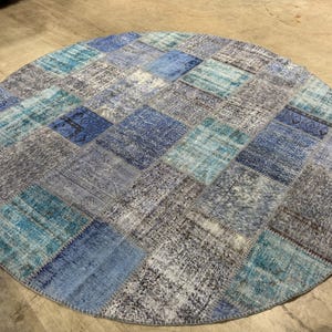 May include: A round area rug featuring a patchwork design composed of various rectangular segments in shades of blue, teal, and grey. Each segment displays distinct faded patterns and textures, stitched together to form a cohesive circular shape.