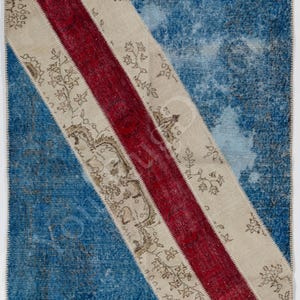 May include: A rectangular area rug featuring a patchwork design with a large blue section on the left and right, separated by a diagonal band of cream fabric with brown floral patterns and a central stripe of deep red fabric with a textured finish.